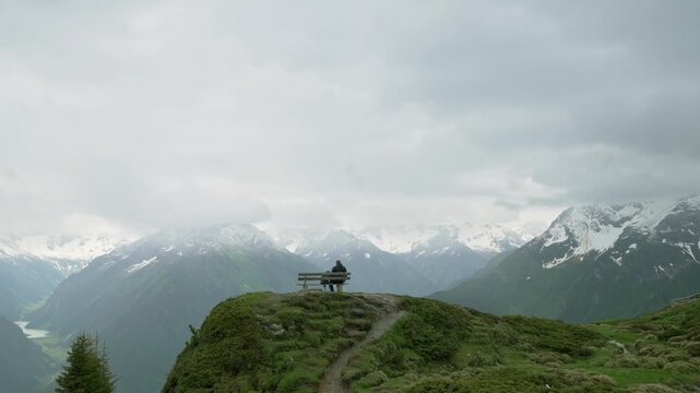 A solitary hiker stands atop a grassy hill in Penken, Austria, gazing at the expansive view of snow-capped mountains and valleys under a cloudy sky, epitomizing the beauty of alpine exploration.