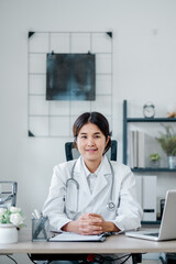 A professional female doctor in a white coat sits at her desk in a modern medical office, ready to assist patients.