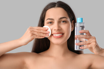 Beautiful young happy woman with cotton pad and bottle of micellar water on grey background