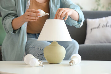 Young woman screwing light bulb into lamp on table at home, closeup