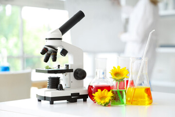 Female scientist with flasks of samples, sunflowers and microscope on table in laboratory, closeup
