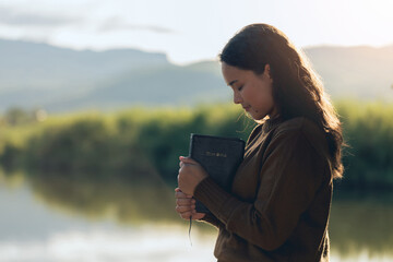 Naklejka premium Christian woman praying on the holy bible in a field during a beautiful sunset. woman standing with closed eyes with the Bible in his hands, Concept for faith, spirituality, and religion. Wishing.