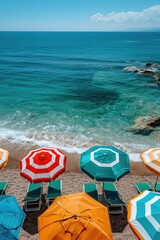 Obraz premium Brightly colored umbrellas are arranged on a sandy beach against the backdrop of clear turquoise water and rocky outcrops. A picturesque and inviting seaside scene.