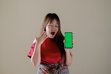 A Southeast Asian woman wearing luxury traditional Indonesian batik outfit is showcasing a digital product and looking at a green screen smartphone with shocked face expression on isolated background