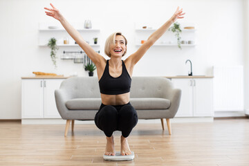 Happy woman celebrating successful weight loss on scale in bright living room. Fitness goals and...