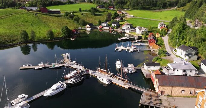 Aerial dolly over idyllic European wooden harbor with boats docked at piers next to homes on grassy field. Clouds in sky reflect on water.