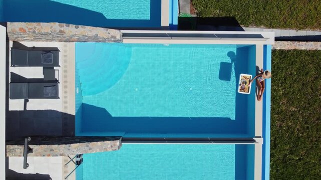 Down view of woman sitting by pool and sunbathing while enjoying food and drink. Single adult enjoying summer vacation relaxation.