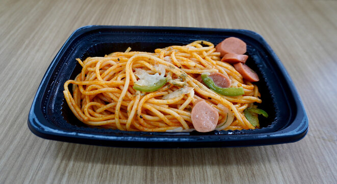 Cheap convinience store spaghetti with tomato sauce and sausage meal in black plastic plate container isolated on horizontal wooden table surface background.