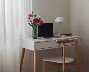 A white table with a laptop, a desk lamp, a notebook and a vase of flowers in the interior of the apartment. Minimalism style