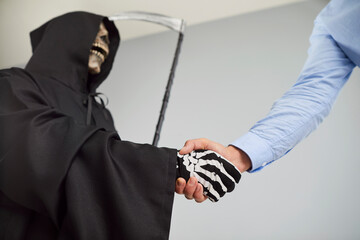 DDevil in skull mask and Halloween grim reaper death costume with black cloak and hood holding scythe shaking hands with a man employee in shirt making deal with devil on gray background.