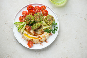 White plate with hummus, falafel and fresh vegetables, flat lay on a light-beige stone background, horizontal shot with space