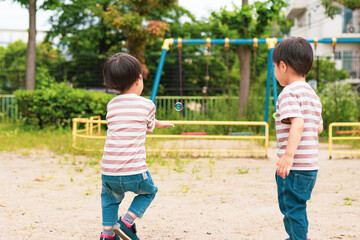 Obraz premium 晴れた昼下がりの公園で、遊具を見つけて向かっている日本の双子兄弟。奥にはブランコがあり木が生い茂っている。