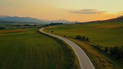 An aerial view of a winding road through rolling green hills at sunset. The sky is a soft orange and yellow, and the mountains in the distance are hazy.