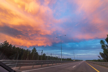 A breathtaking display of colorful clouds illuminates the sky above a quiet stretch of highway lined with tall trees during a tranquil evening.