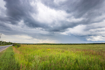 A paved road runs through a field of tall green grass, with dark storm clouds filling the sky above. A hint of sunlight peeks through the clouds in the distance..
