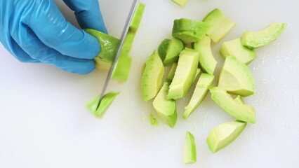Half of green avocado cutting close up on white kitchen board. Chef hands in blue gloves chop fresh ripe vegetable, iron knife. Cooking, preparing ingredients in professional restaurant. Top down view