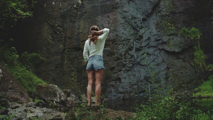 Naklejka premium Woman admires a hidden waterfall in a lush tropical forest, touching her hair in wonder. Perfect for summer adventures in french polynesia's moorea