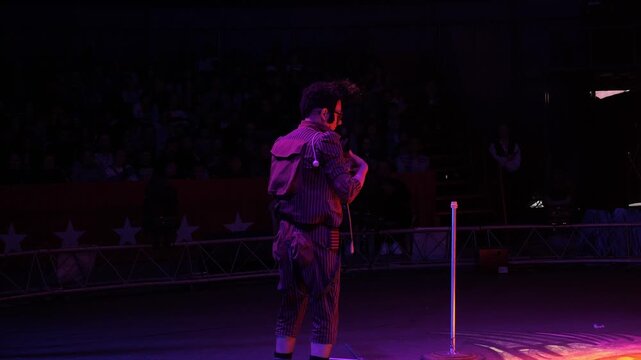 A clown with a funny hairstyle entertains guests in the circus arena in the light of colorful spotlights and smoke. He takes something out of his pocket. Circus performance.