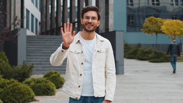 Hello. Indian man smiling friendly at camera, waving hands gesturing hi, greeting or goodbye, welcoming with hospitable expression outdoors. Happy Arabian Hindu guy standing in urban city town street