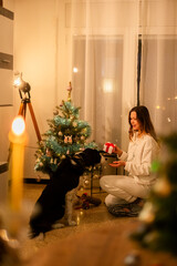Fotograf&iacute;a en vertical de un sal&oacute;n donde hay una chica en pijama blanco con un regalo en la mano y con la otra llama a su perro estilo Border Collie.