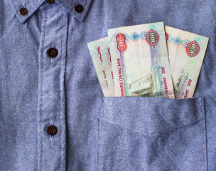 One hundred dirhams poking out from a pocket of an office style blue color shirt with dark brown buttons. Paper money, closeup