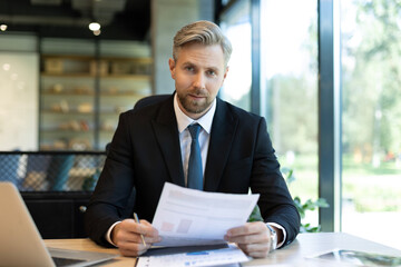 Fototapeta premium Mature businessman with beard working with documents, contracts and bills sitting at table using laptop at work in modern bright office with large windows