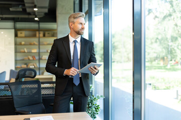 Portrait of smiling middle aged businessman standing with digital tablet at corporate office