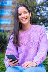 Front view of a laughing latin teenage girl using a social media application with her mobile phone, leaning on the wall looking at camera. University life concept.Vertical