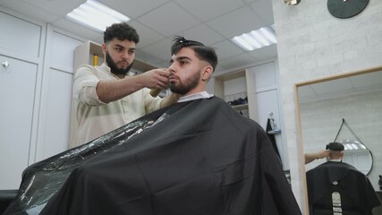 The process of working in a modern barber shop for men. Barber cuts a client with a beard on the background of mirrors and haircut tools.