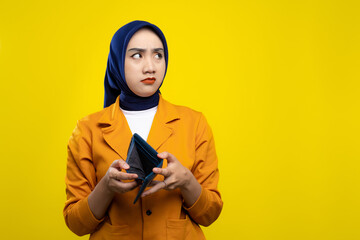 Sad young Asian woman holding empty wallet and looking aside at copy space with worried expression isolated on yellow background