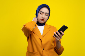 Tired young Asian woman using smartphone and feeling pain in her neck isolated on yellow background