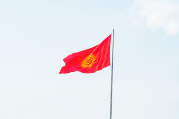 Kyrgyzstan national flag featuring a red background with a yellow sun and a traditional tunduk symbol. Symbol of unity and heritage. Waving prominently on a flagpole against a clear, blue sky.