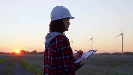 Woman engineer is taking notes on a clipboard on a field with wind turbines, as the sun sets in evening. Clean energy and engineering