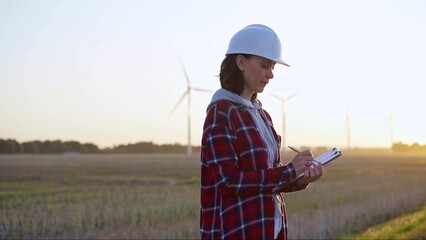 Adult woman engineer taking notes on a clipboard on a field with wind turbines, as the sun sets