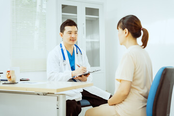 Obraz premium A male Asian pediatrician in a white lab coat sits at his desk, warmly greeting a middle-aged pregnant woman for a prenatal consultation, offering care and support in a clinical setting.
