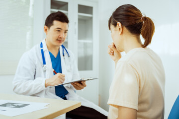 A male Asian pediatrician in a white lab coat sits at his desk, warmly greeting a middle-aged pregnant woman for a prenatal consultation, offering care and support in a clinical setting.