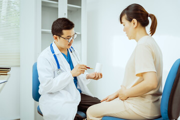 Fototapeta premium A male Asian pediatrician in a white lab coat sits at his desk, warmly greeting a middle-aged pregnant woman for a prenatal consultation, offering care and support in a clinical setting.