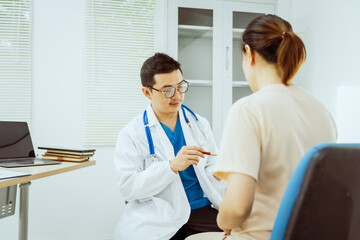 A male Asian pediatrician in a white lab coat sits at his desk, warmly greeting a middle-aged pregnant woman for a prenatal consultation, offering care and support in a clinical setting.