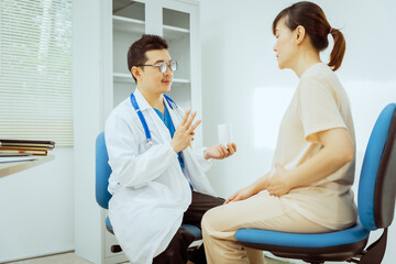 Fototapeta premium A male Asian pediatrician in a white lab coat sits at his desk, warmly greeting a middle-aged pregnant woman for a prenatal consultation, offering care and support in a clinical setting.
