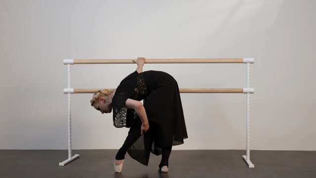 A beautiful ballerina in a black dress dances in a white sunny room with a black floor next to a ballet barre.