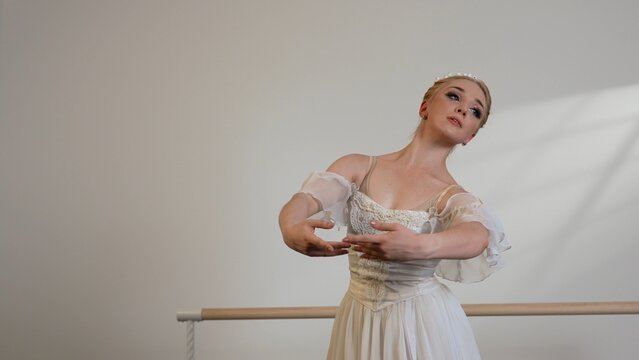 A beautiful ballerina in a white dress dances in a white sunny room next to a ballet barre.