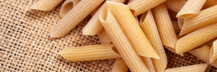 Raw Italian penne pasta on rough burlap close up. Web banner element. Uncooked traditional food. Macro shot