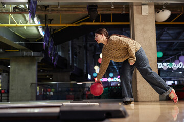 Woman in cozy sweater and jeans bowling in modern, brightly lit alley with vibrant lights and hanging screens creating dynamic atmosphere