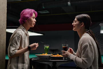 Two friends are enjoying conversation at a bar, both holding drinks with snacks on table. The scene features two people engaged in a relaxed social setting