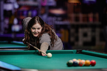 Woman with focused expression playing billiards, bending over billiard table and preparing for...