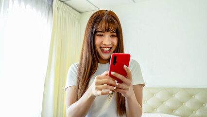 Happy asian young woman sit on bed at home typing on mobile phone receive great news celebrating...