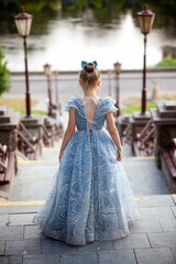 Fototapeta premium A beautiful little blonde princess in a blue medieval dress. The lovely queen poses on the stone steps on a warm summer day. Rear view.