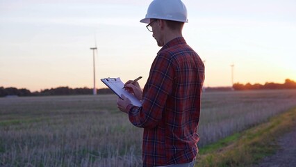 Man engineer wearing a white protective helmet is taking notes with a clipboard in a field with wind turbines, as the sun sets. Clean energy and engineering concept