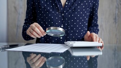 Auditor woman analyzing financial documents with magnifying glass and calculator at her office desk in formal dark blue blouse with polka dots. Business people concept