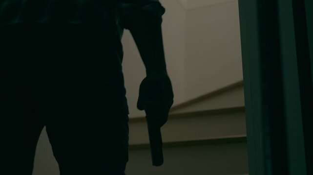 Slow motion of a man in a striped shirt, cap and holding a pistol walking up stairs. Concept of burglary, trespassing, murder.